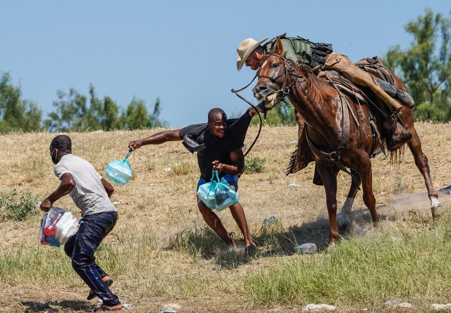 A border-patrol officer on horseback leans over and grabs the shirt of a man on foot who is carrying bundles of food and trying to escape.