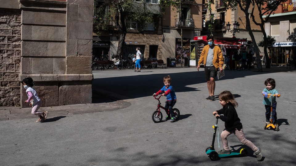 A man in a mask oversees a group of children playing on bikes and scooters in a plaza outside.