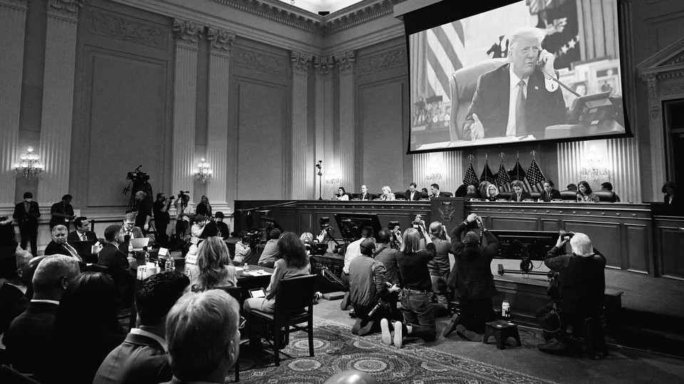 Black-and-white photo of January 6 hearings; photographers kneel on the floor in one part of the room, aiming their cameras at a screen showing Donald Trump on a phone