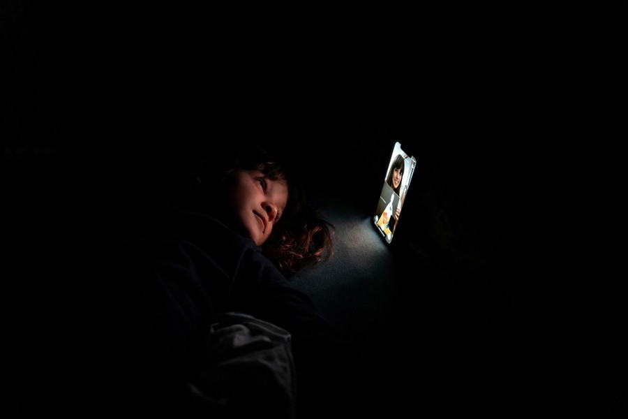 A young girl lies in front of a smartphone, her face lit by its screen, in a dark room.