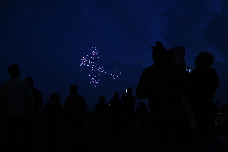 People look up into the night sky, watching a drone light show, as drones form the outline of a WWII-era fighter plane.