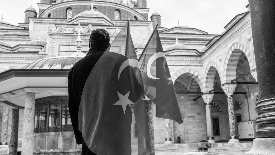 A black-and-white photo taken from behind a man looking at a mosque while holding two Turkish flags in his right hand.
