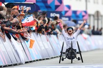 An athlete in a racing wheelchair raises their arms in celebration as they pass a crowd at a finish line.