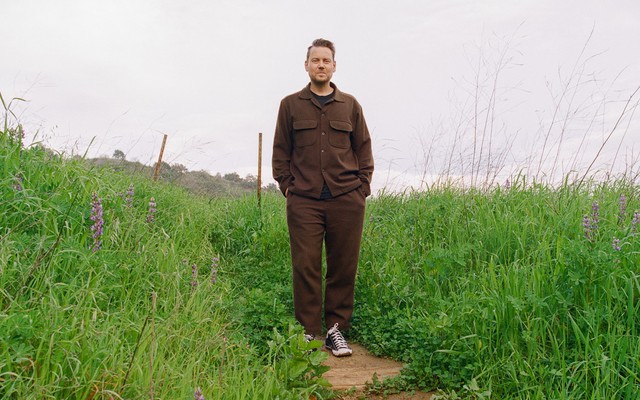Color photo of man in brown shirt and pants standing with hands in pockets on trail surrounded by green grass and wildflowers.