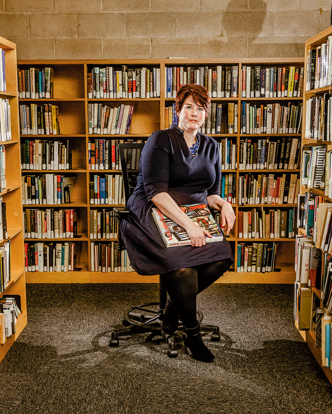 A photograph of a woman sitting along lines of books