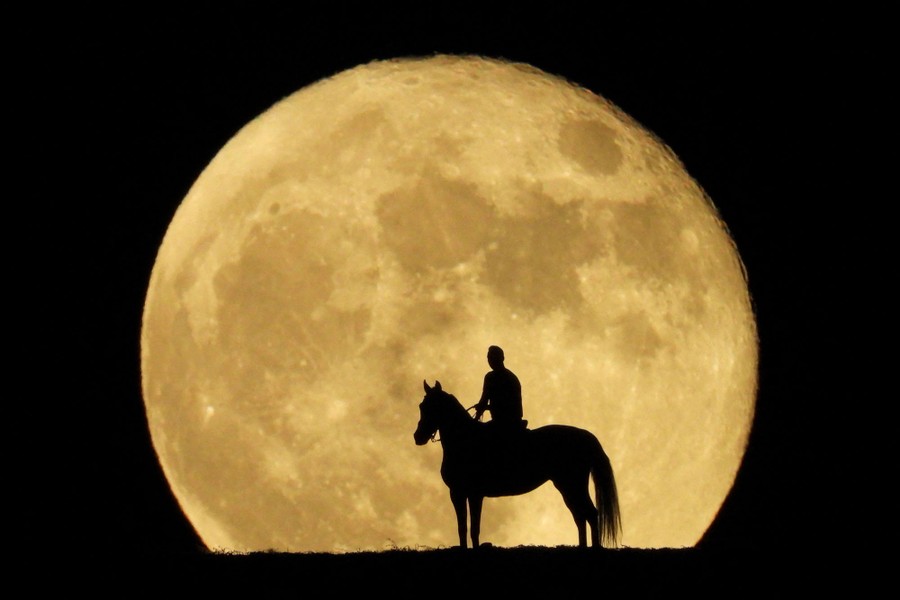A person atop a horse is silhouetted in front of a large full moon.