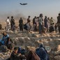 Women and children sit on dusty rocks with men standing behind, facing distant airport walls and watching a military plane taking off
