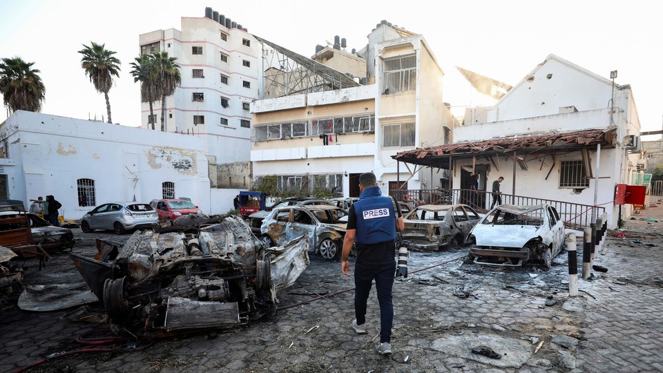 A person wearing a blue PRESS vest and carrying a large camera walking into a burned-out parking lot in Gaza.