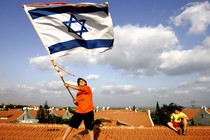 A person waving a large Israeli flag while standing on a roof with a big sky in the background