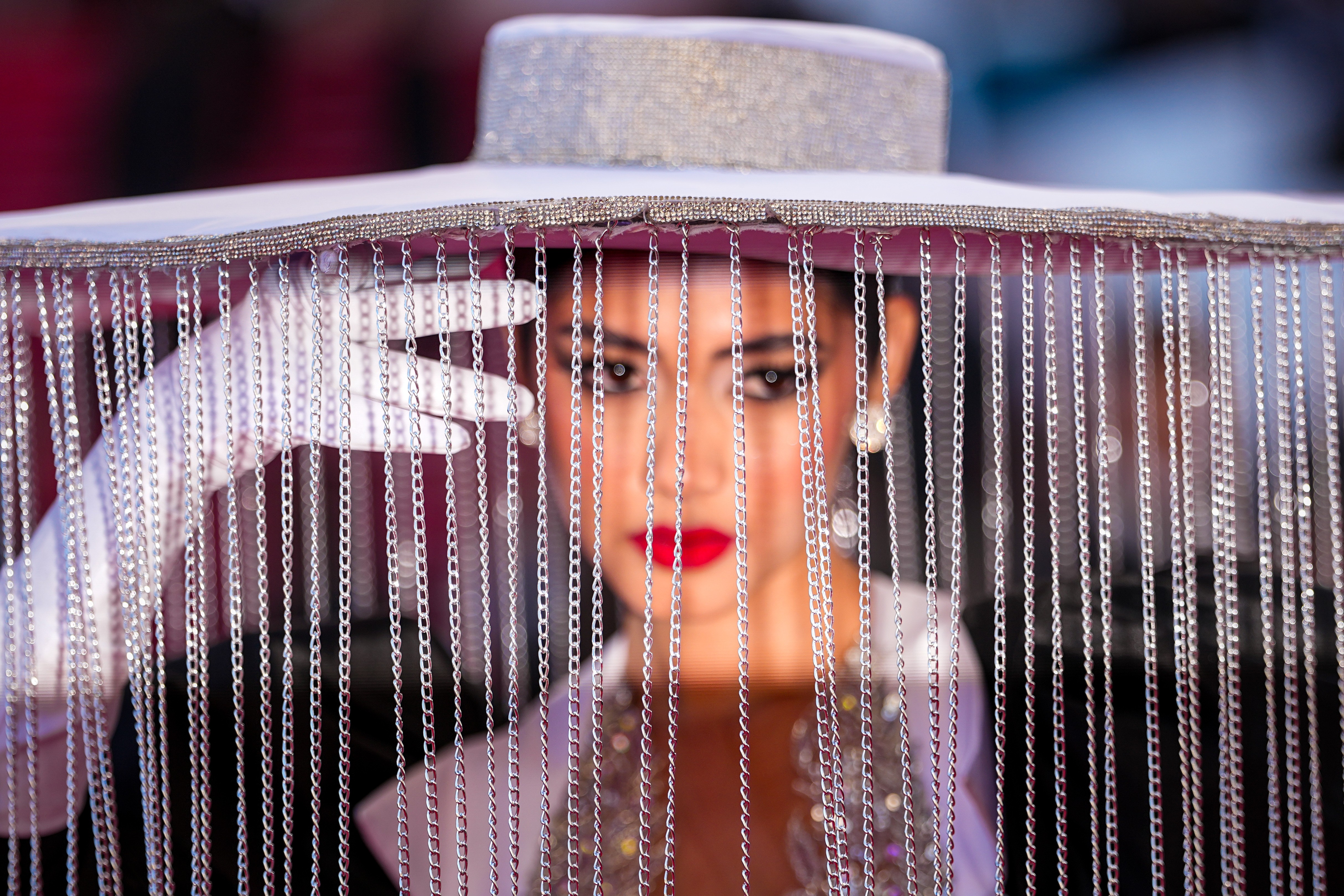 A model poses for photographers, wearing a broad-rimmed hat with thin chains hanging from the brim, forming a veil.