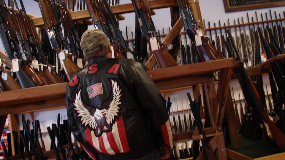 A man in a jacket featuring a bald eagle and the American flag stands in front a collection of rifles.