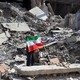 Photograph of a man holding up an Iranian flag atop the wreckage of a bombed building
