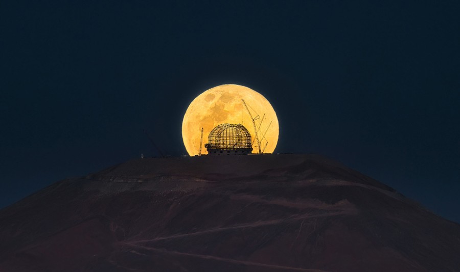 A full moon rises behind a partially-built dome that will house a large telescope.