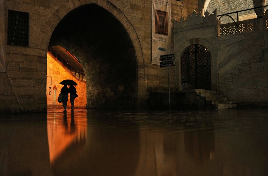 Two people walk with an umbrella through an arch, reflected by rainwater on the ground.