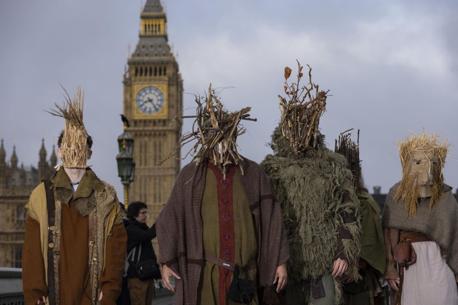 Four people in traditional costumes, with masks made of sticks and straw, stand in front of the Palace of Westminster, in London.