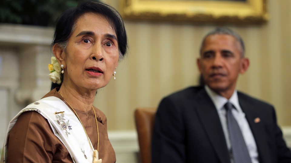 Aung San Suu Kyi looks at the camera while Barack Obama looks at her in the background. They are sitting in the Oval Office.