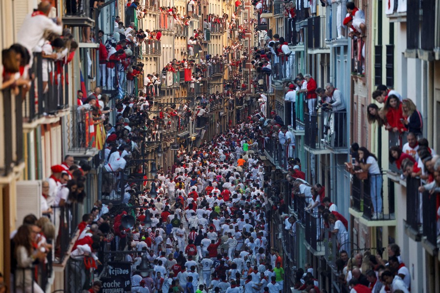 People watch from balconies along a road lined with tall buildings as hundreds run in the street below.