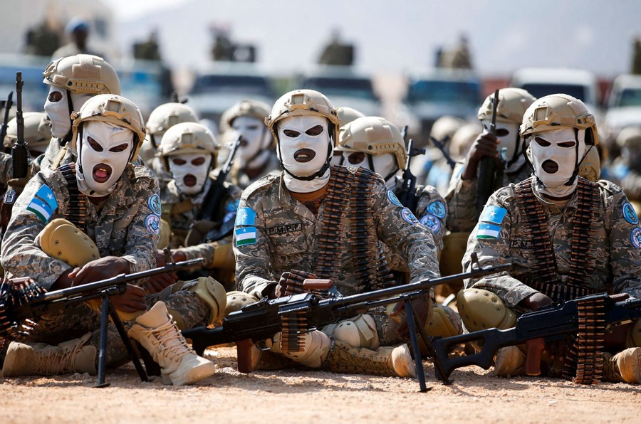 Soldiers in uniform, and wearing white balaclavas, sit together, part of a parade.