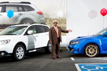 A car dealer in a brown suit holding his arm outstretched toward a blue Subaru with a red balloon tied to it