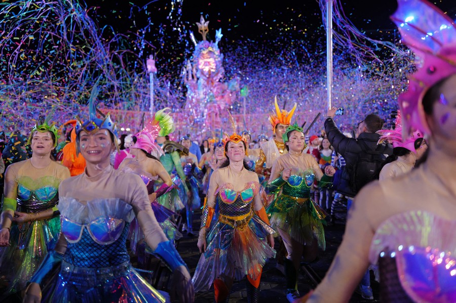Costumed performers march in a parade, beneath streamers and confetti.