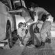 Una fotografía en blanco y negro de una mujer con una camiseta y jeans apoyada con ambas manos contra una camioneta blanca,mientras una mujer uniformada la inspecciona y una niña pequeña llora, por la noche.
