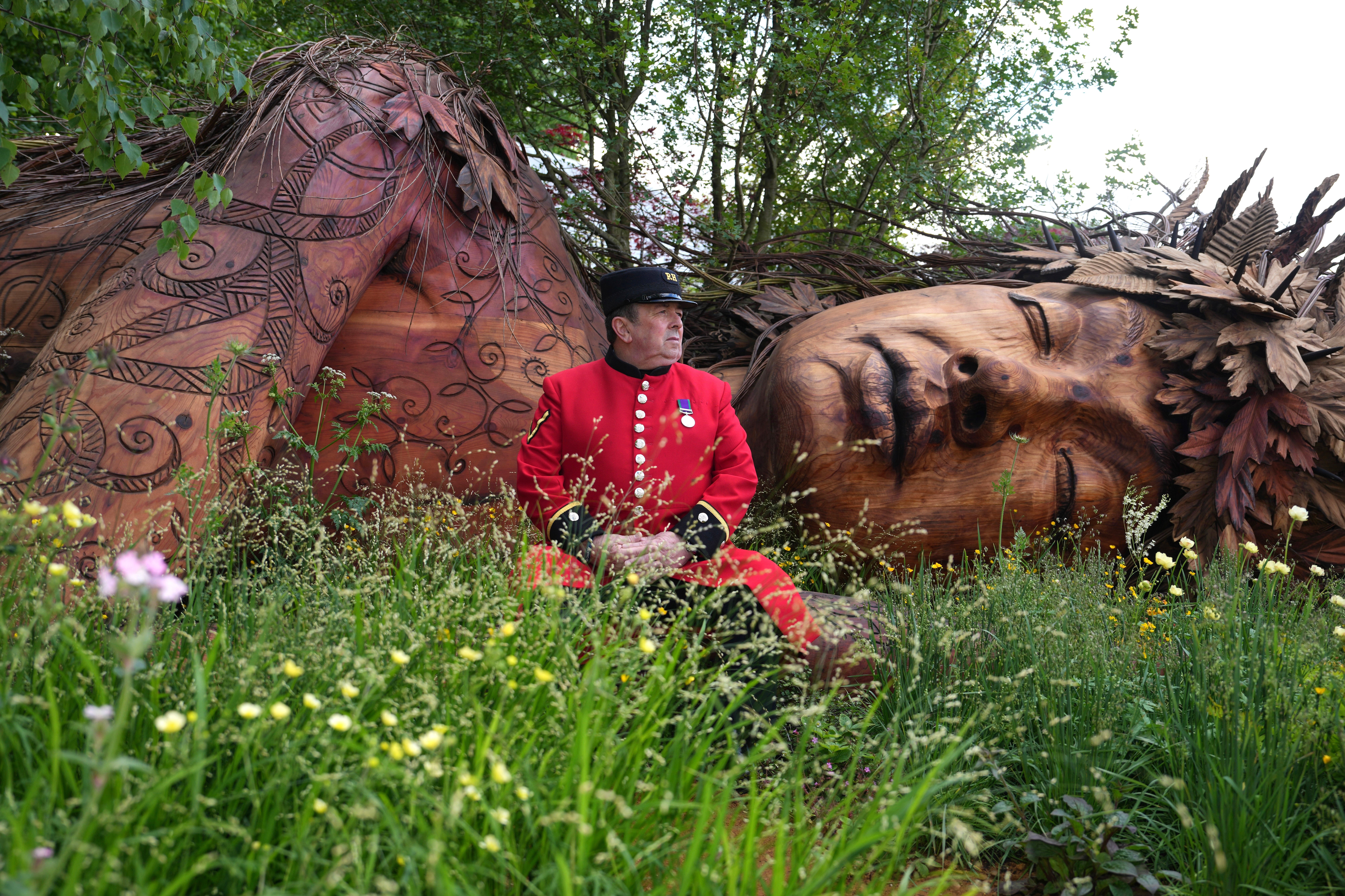 An older man in a dress military uniform sits beside a large wooden sculpture in a garden.