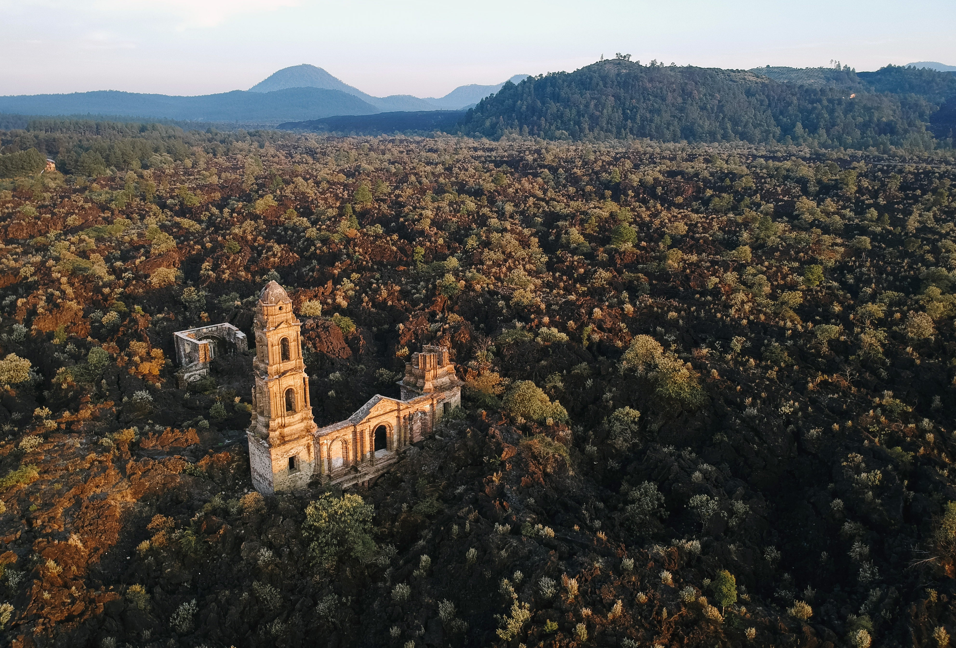 Aerial view of the bell tower of the ruined church, standing in a wide field of overgrown volcanic debris.