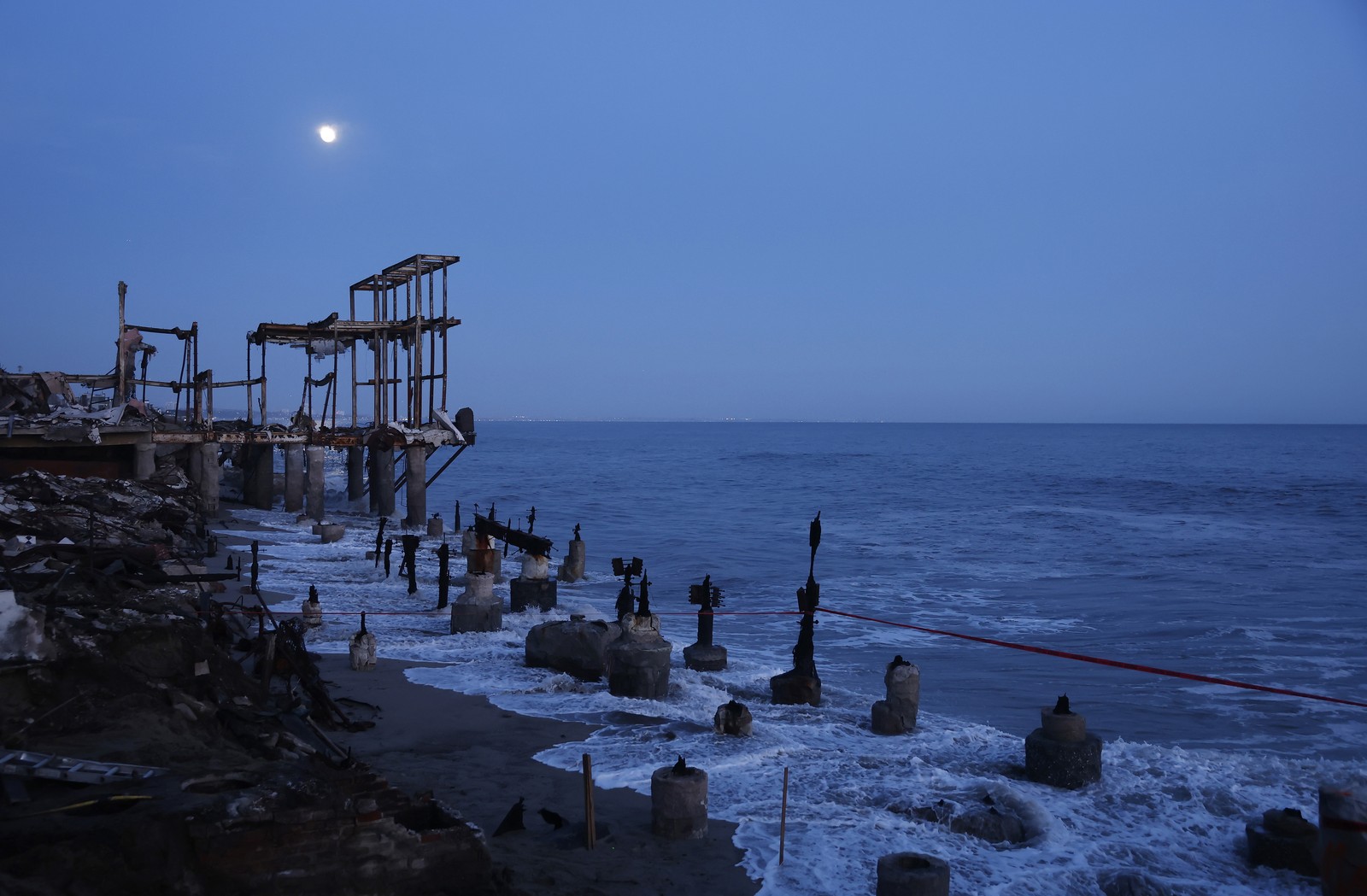 The moon rises above the ruins of a burned-out building, on a shoreline.