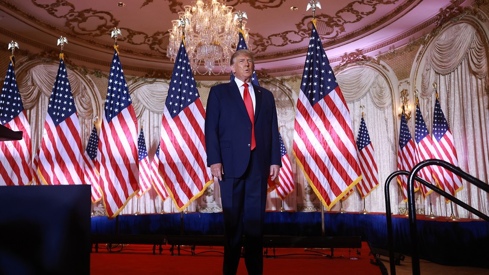 Donald Trump stands on a red carpeted stage, flanked by American flags.
