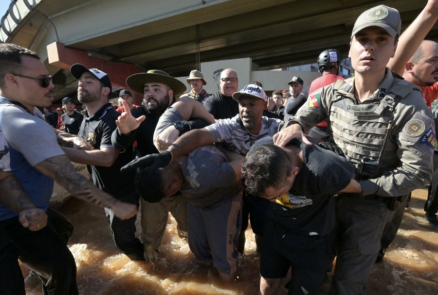 Police officers escort two handcuffed people through knee-deep floodwater as a crowd gathers around them.