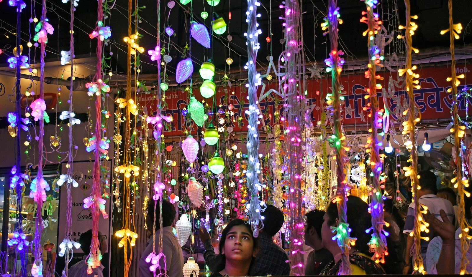 A girl in a marketplace looks up at colorful strings of electric lights.