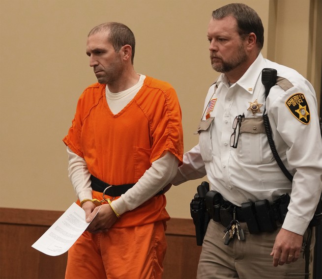 photo of balding man in handcuffs and orange prison clothes being escorted by a uniformed sheriff 