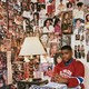 detail of photo of teen boy in red and blue jersey sitting with hands clasped and leaning against wall covered in cut-out magazine pages and sports posters