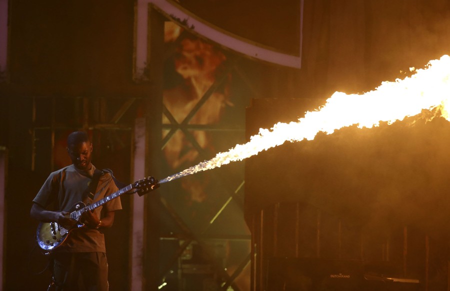 A man plays a guitar with a flamethrower attachment blasting a jet of fire from its neck.