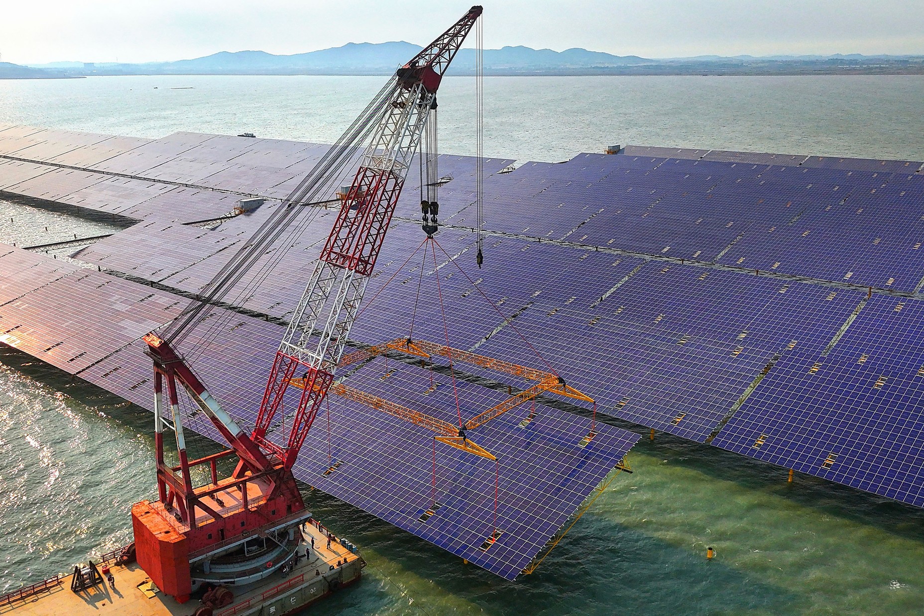 A crane on a floating barge positions a large solar panel for installation.