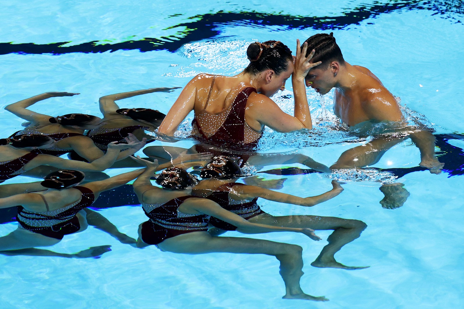 An artistic swim team prepares to lift one of their members out of the water, while she interacts with another team member nearby.