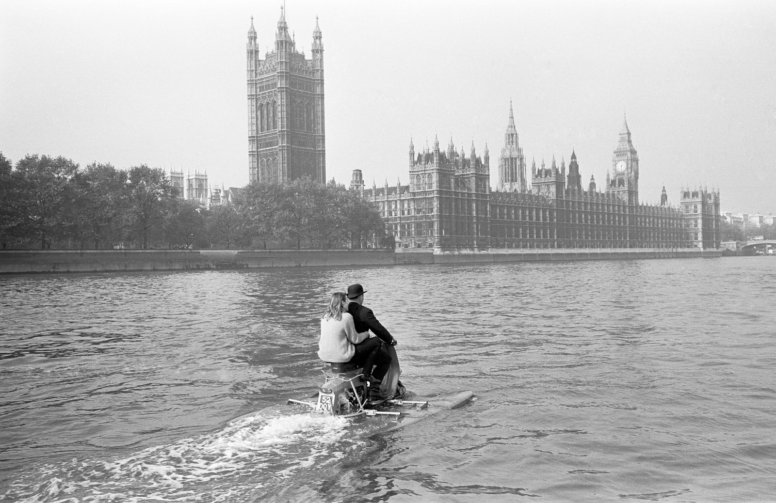 A man and his daughter ride on a scooter that is floating and paddling down the River Thames, with the Palace of Westminster visible in the background.
