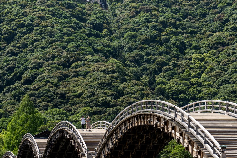 People walk across a set of wooden-arch bridges near a wooded hillside.