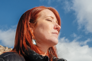 photo of red-haired woman with eyes closed wearing black jacket in sunlight with mountain and blue sky in background