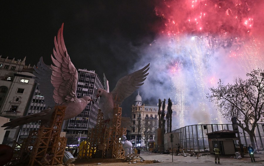 Fireworks explode above a city square, with two large dove statues in the foreground.