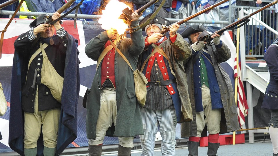 Six men dressed in reenactment costumes firing muskets. An audience in football jerseys and rain ponchos sit behind them.