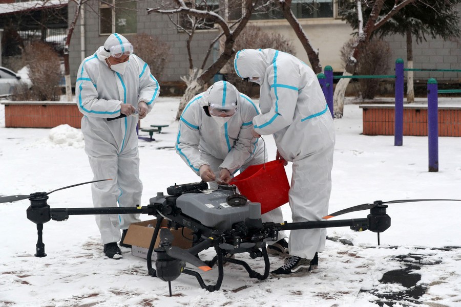 Workers in protective suits use a red bucket to fill a tank in a drone.