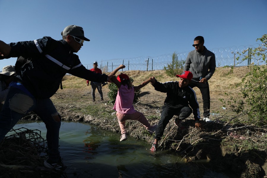 Several people reach out to lift a small child across a small section of a river.