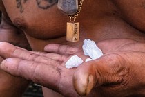 Close-up view of 3 bright-white crystal rocks resting in a man's palm in front of his bare tattooed chest and necklaces, one with a rectangular pendant with the word "POWER"