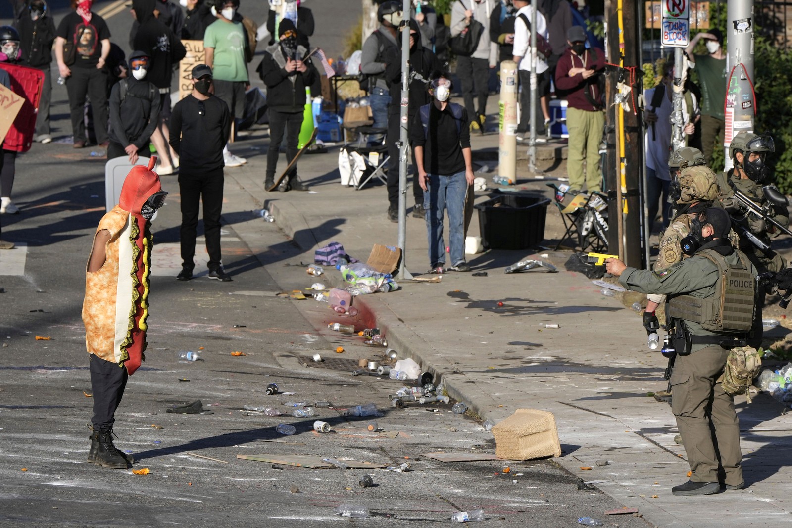 A law enforcement officer points a taser at a person wearing a hot dog costume during a protest.