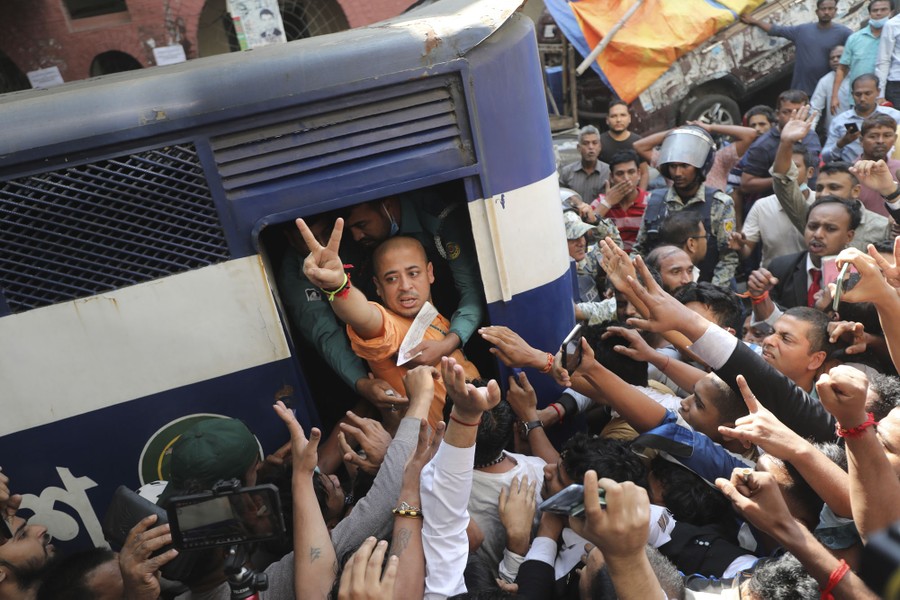 A crowd crushes in around a prison van as a man in the van's doorway raises a hand, making a "V" with two fingers.