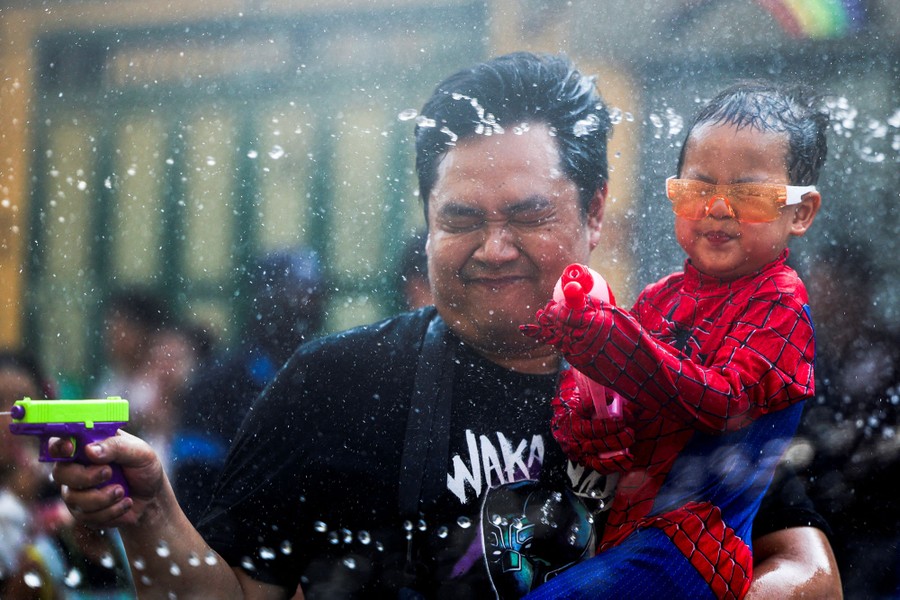 A person holds a child wearing a Spider-Man costume, both of them soaked and spraying water pistols.