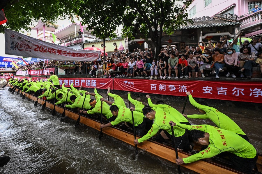 A crowd of spectators sits along the edge of a canal, watching as a very long dragon boat passes by, being rowed by more than a dozen people.