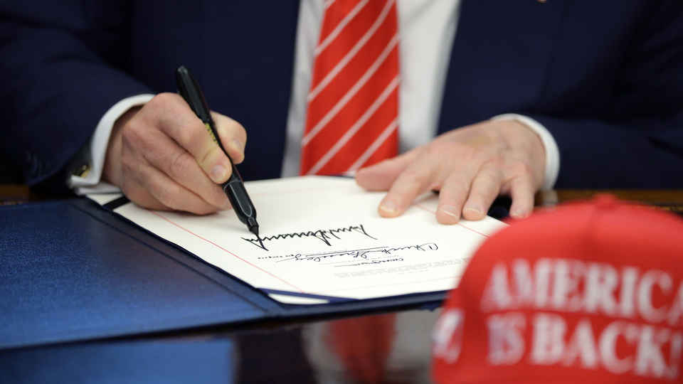 A close-up of Trump's hand and pen as he signs a document while wearing a striped red and white tie