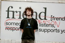 A man with curly hair stands in front of a billboard at a subway station.